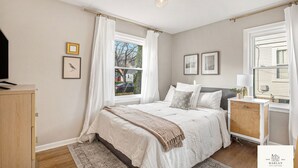 A bedroom with a bed, a nightstand, a dresser, and a window with curtains. - The Harlan City Cottage - Extended Stay Welcome (Grand Rapids)