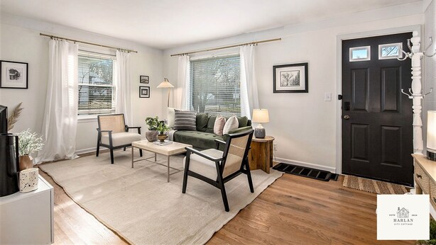 A living room with a sofa, chairs, a coffee table, and a lamp. There is a black door with a coat rack and a window with curtains. - The Harlan City Cottage - Extended Stay Welcome (Grand Rapids)
