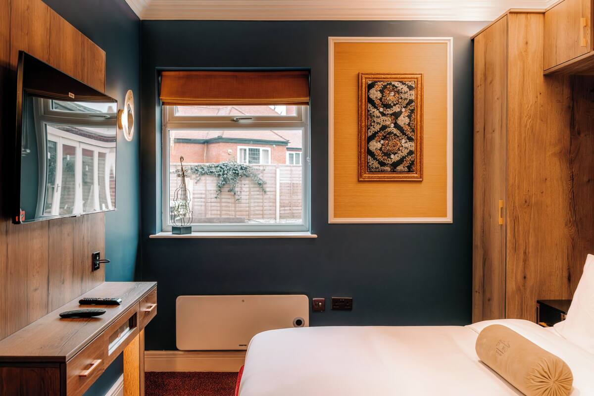 Une chambre d’hôtel comprenant un lit, un bureau avec une chaise, une fenêtre donnant sur des immeubles et une œuvre d’art accrochée au mur.