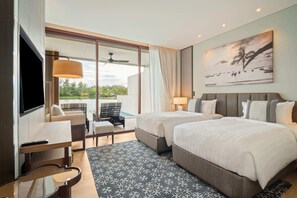 Une chambre d’hôtel avec deux lits, une télévision, un coin repas avec une table et des chaises, et un balcon donnant sur un espace verdoyant.