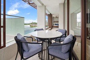 A balcony with a glass railing, a round table with chairs, and a bed with a view of the ocean.