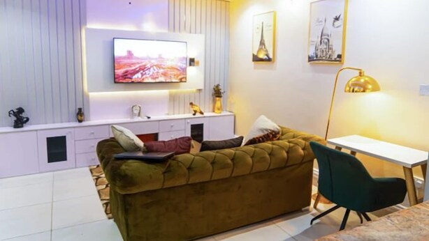 A modern living room with a sofa, a TV mounted on the wall, a desk, and a lamp. - The Panache Luxury Apartment (Lagos)