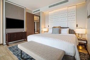 Une chambre d’hôtel moderne avec un grand lit, un téléviseur à écran plat fixé au mur, une armoire en bois, deux lampes de chevet et un tapis à motifs.