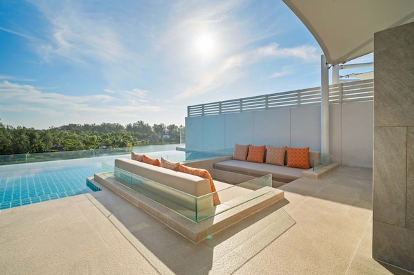 A modern outdoor pool area with a glass balustrade, beige sofas with orange cushions, and a clear view of the surrounding landscape.