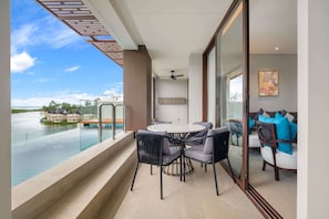 A balcony with a glass railing overlooking a body of water, a round table with chairs, and a seating area with cushions.