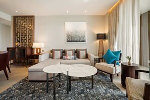 A modern hotel room with a sofa, two round tables, a patterned rug, a decorative wall panel, a flat-screen TV, and a large window with curtains.