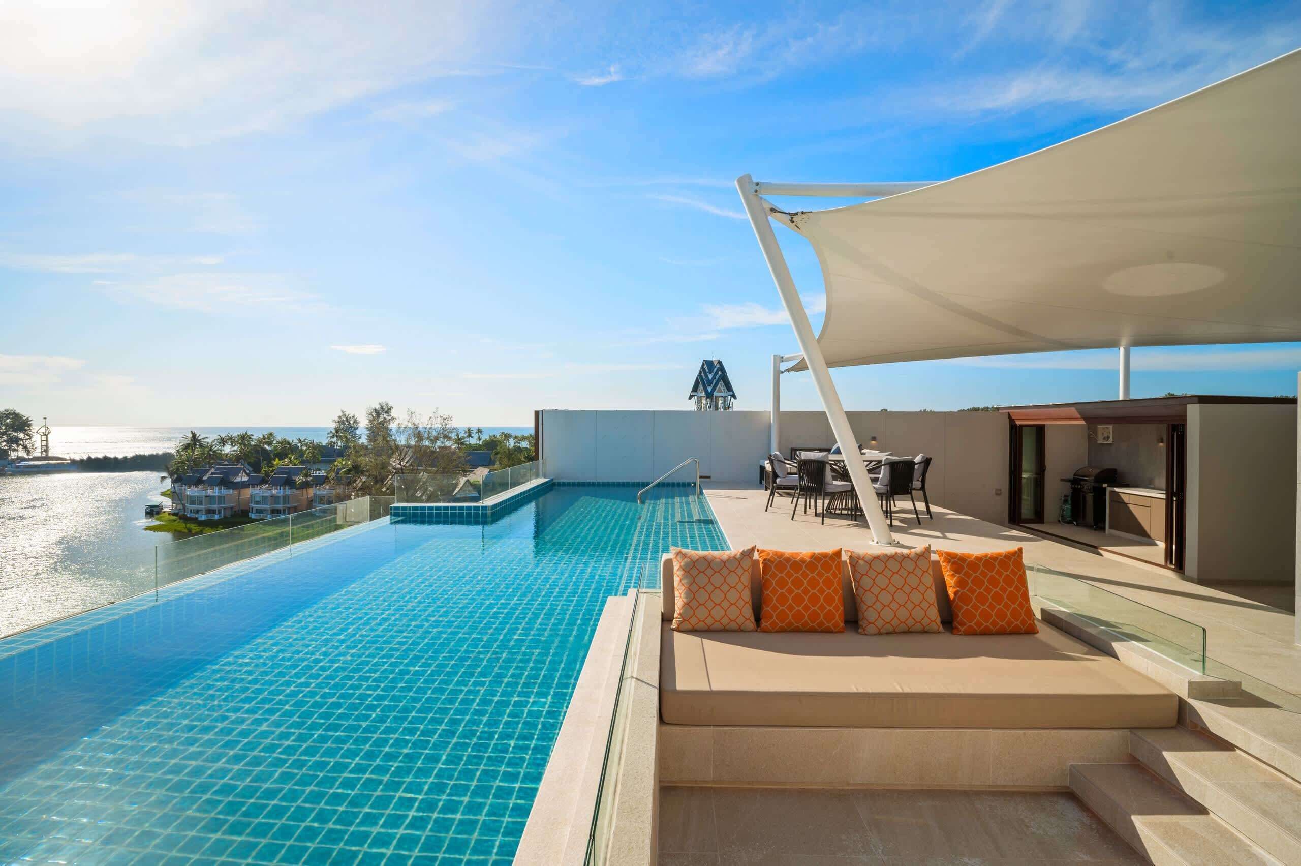 A rooftop infinity pool with a view of the sea, a seating area with orange patterned cushions, and a covered outdoor dining space.