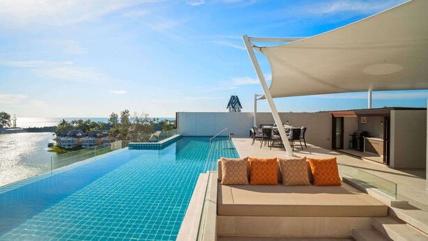 A rooftop infinity pool with a view of the sea, a seating area with orange patterned cushions, and a covered outdoor dining space.