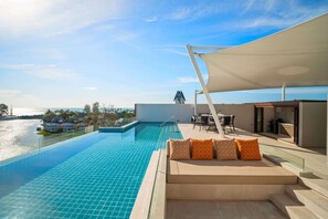 Una piscina infinita en la azotea con vistas al mar, un área de descanso con cojines de estampado naranja y un espacio de comedor al aire libre cubierto.