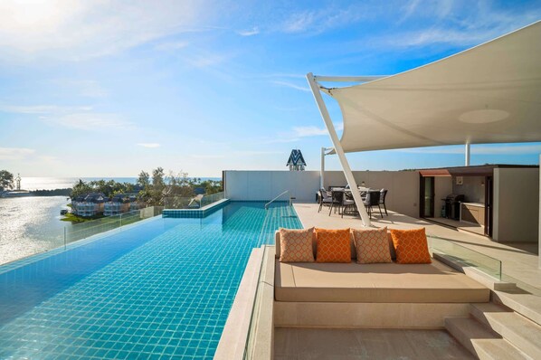 A rooftop infinity pool with a view of the sea, a seating area with orange patterned cushions, and a covered outdoor dining space.