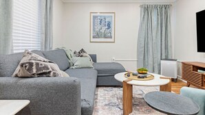 A modern living room with a grey sofa, a round coffee table, and a flat-screen TV on a wooden stand. - Soak and Play: Hot Tub + Family Arcade Fun (Euclid)