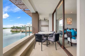 A balcony with a glass railing overlooking a body of water, a round table with chairs, and a seating area with cushions.