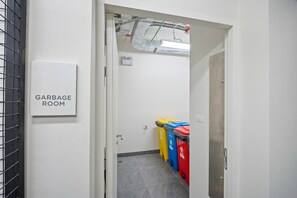 Un couloir menant à une pièce où se trouvent des poubelles, avec une pancarte, une porte et trois conteneurs à ordures.