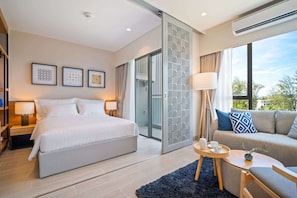 Een moderne hotelkamer met een groot bed, een bank, een klein tafeltje met dienblad en een groot raam met uitzicht op groen.