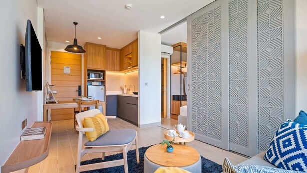 A modern living room with a sofa, a chair, a coffee table, and a TV. There is a kitchen area with wooden cabinets and a microwave.