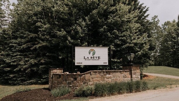 A sign for 'La Bête Golf Club' with a stone wall and greenery.