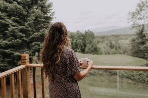 A person standing on a wooden deck holding a cup, overlooking a grassy area with trees and mountains in the distance.