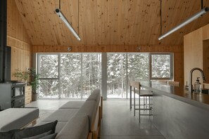 A modern interior with a wooden ceiling, large windows, a sofa, a bar area, and a kitchen. - Woodland Getaway | Hot Tub • Sauna • Hike • Fire (Chertsey)
