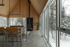 A cabin interior with a dining area, a fireplace, and large windows offering a snowy forest view. - Woodland Getaway | Hot Tub • Sauna • Hike • Fire (Chertsey)