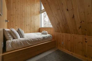 Une chambre avec un plafond en pente, des murs en bois, un lit avec des oreillers, une fenêtre et un petit banc intégré.
