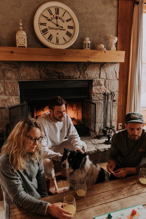 A family around a fireplace with a clock above it, a dog, and a table with drinks.