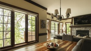 Un amplio salón con una mesa de comedor de madera, una chimenea y grandes ventanales que dan a vistas de árboles y un edificio.