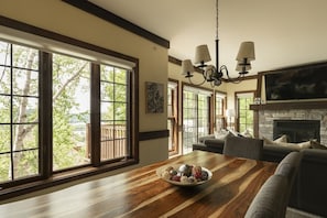 Un amplio salón con una mesa de comedor de madera, una chimenea y grandes ventanales que dan a vistas de árboles y un edificio.