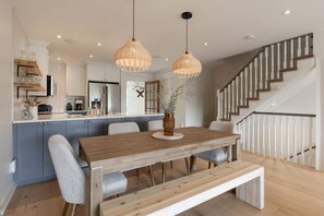 A modern kitchen with a wooden dining table, grey chairs, and pendant lighting.