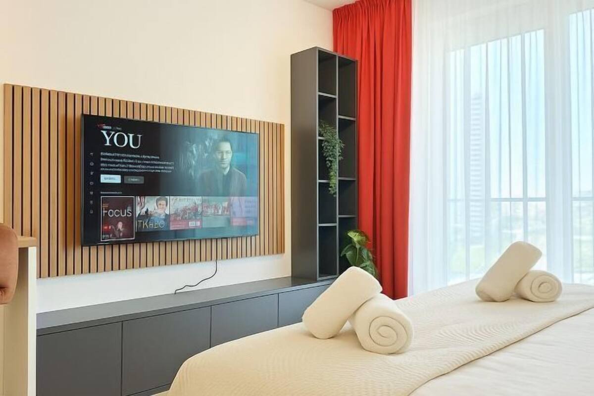 Ein modernes Hotelzimmer mit großem Flachbildfernseher an einer Wand aus Holzpaneelen, einem Bett mit weißen Leinen, einem roten Vorhang und einer Pflanze in einem schwarzen Regal.