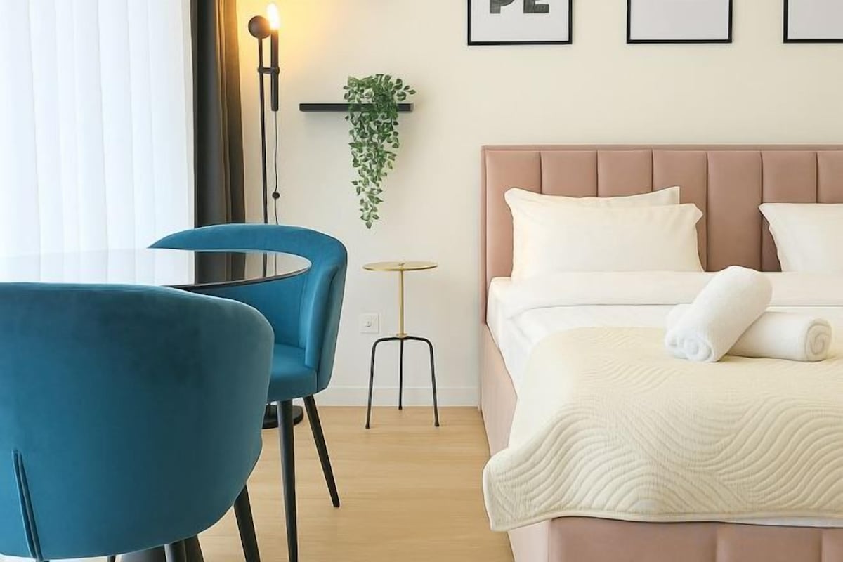 A hotel room with a bed, a bedside table, a chair, and a hanging plant.