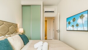 A hotel room with a bed, a headboard, a wall-mounted air conditioner, a door leading to a hallway, and a painting of palm trees. - Waterfront Collection Apartments (Belgrade)