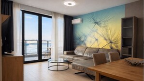 A modern living room with a sofa, a coffee table, a wooden dining table, and a bookshelf. There is a large window with a view of the outdoors. - Waterfront Collection Apartments (Belgrade)