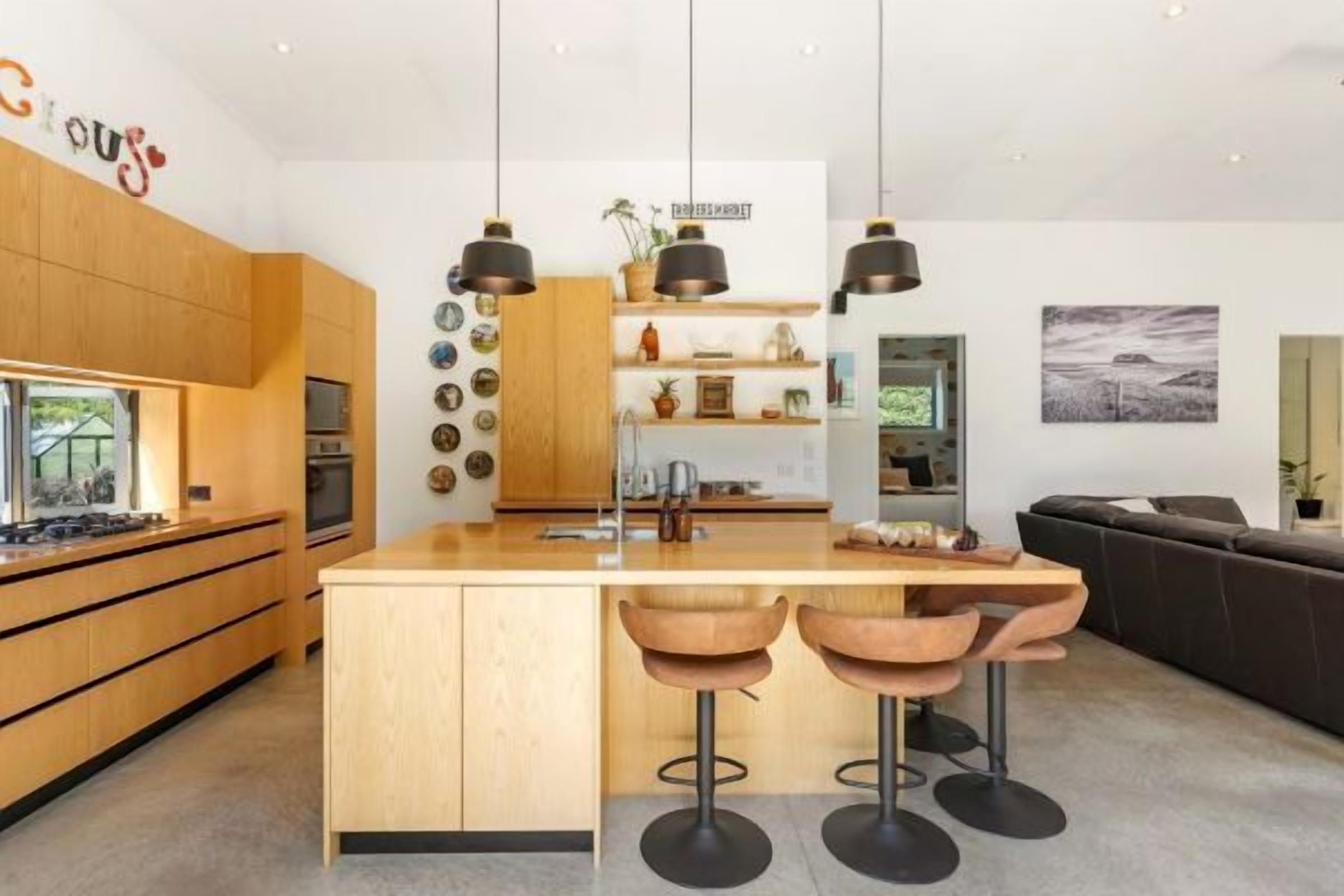 Eine moderne Küche mit Holzschränken, einer zentralen Kochinsel mit Hockern, Pendelleuchten und Blick auf den Wohnbereich mit Sofa und einem Bild an der Wand.
