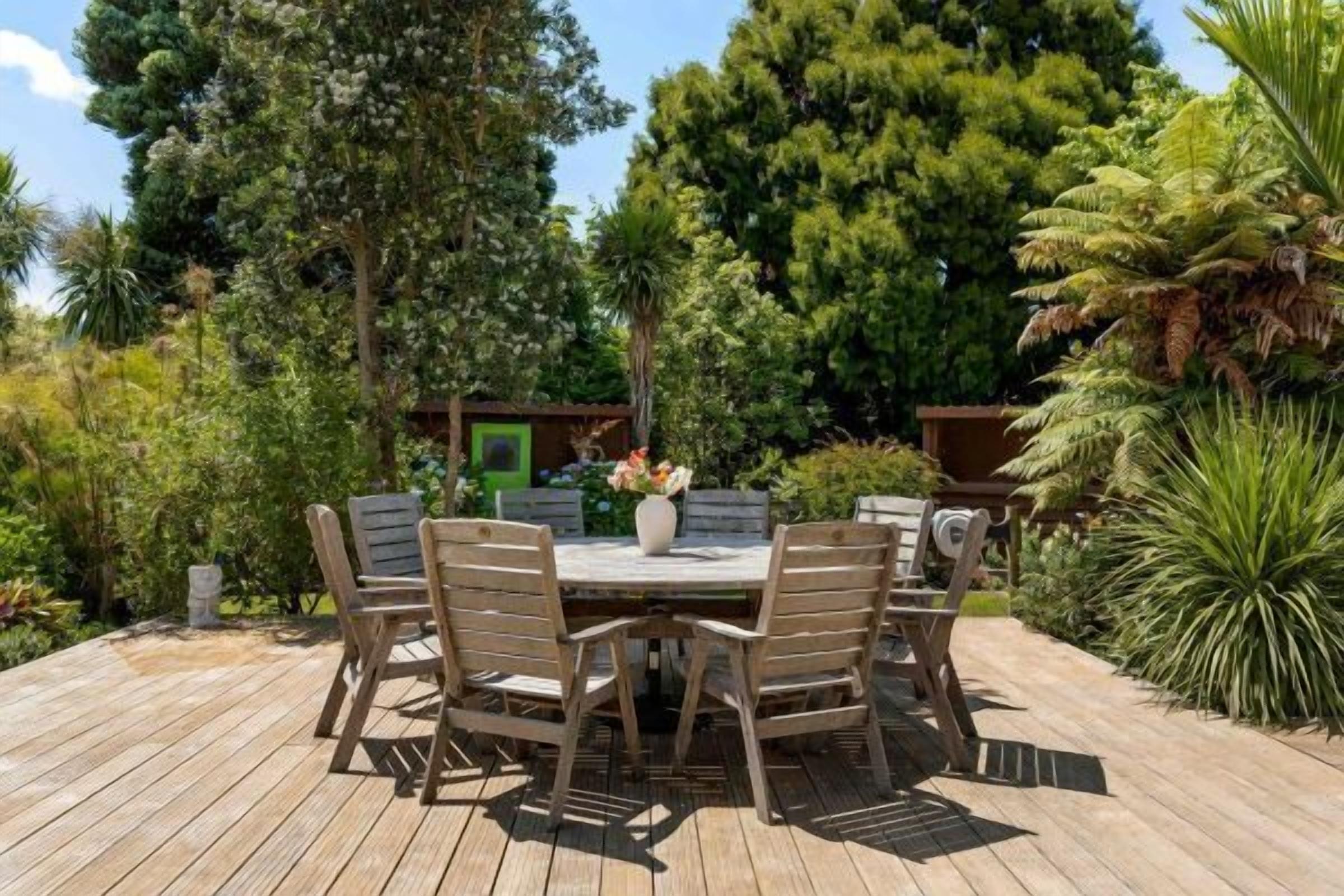 Een houten terras met een ronde tafel en stoelen, omringd door weelderig groen en hoge bomen.