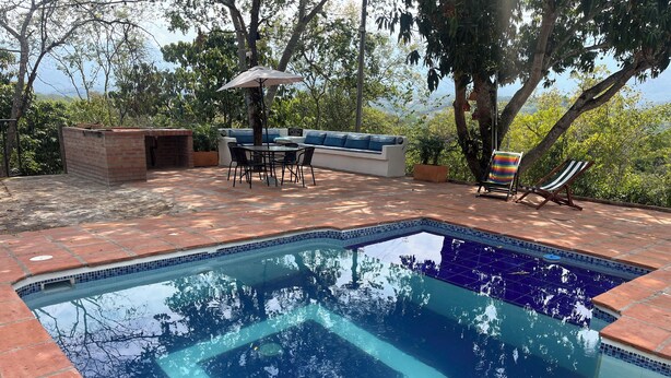 Una piscina rectangular amb rajoles blaves, envoltada d'un pati de maó. Hi ha una taula i caires sota un para-sol, i una cadira relaxant amb coixí ratllat.