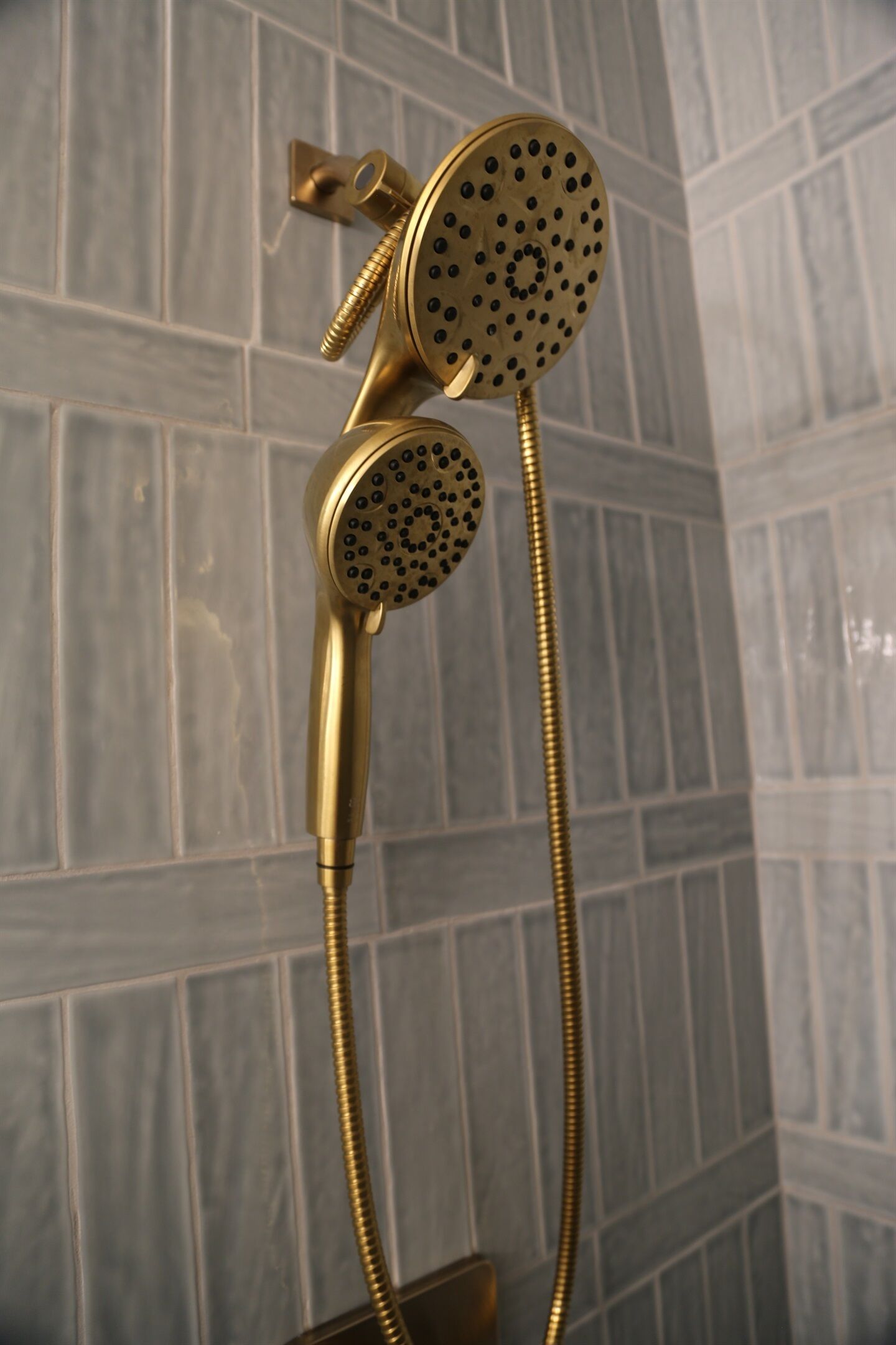 A gold-colored showerhead with a flexible hose, mounted on a wall with a textured tile pattern.