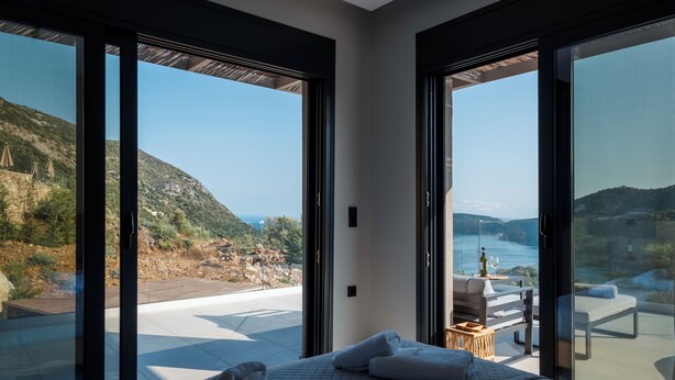 Quarto moderno com uma cama grande, duas janelas lado a lado e vista para uma praia e montanhas.
