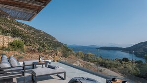 Um terraço com mobiliário para área externa, uma mesa com garrafa e copo, e vista para um lago e montanhas.