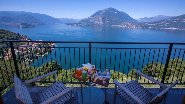 Un balcone con due sedie di legno, un tavolino rotondo e un vassoio con teiera, tazze e un libro. Il balcone si affaccia su un lago con montagne sullo sfondo.