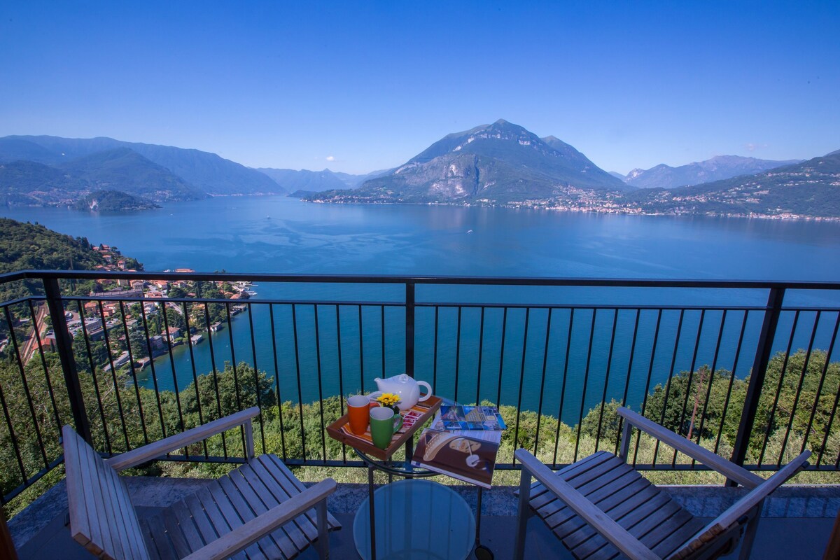 A balcony with two wooden chairs, a small round table, and a tray with a teapot, cups, and a book. The balcony overlooks a lake with mountains in the background.