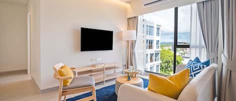 Una habitación de hotel moderna con una televisión de pantalla plana montada en la pared, un sofá con cojines amarillos y azules, una silla de madera, una mesita con una lámpara y un ventanal grande con vistas a la ciudad.