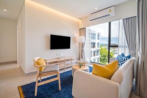 A modern hotel room with a flat-screen TV mounted on the wall, a sofa with yellow and blue cushions, a wooden chair, a small table with a lamp, and a large window offering a city view.