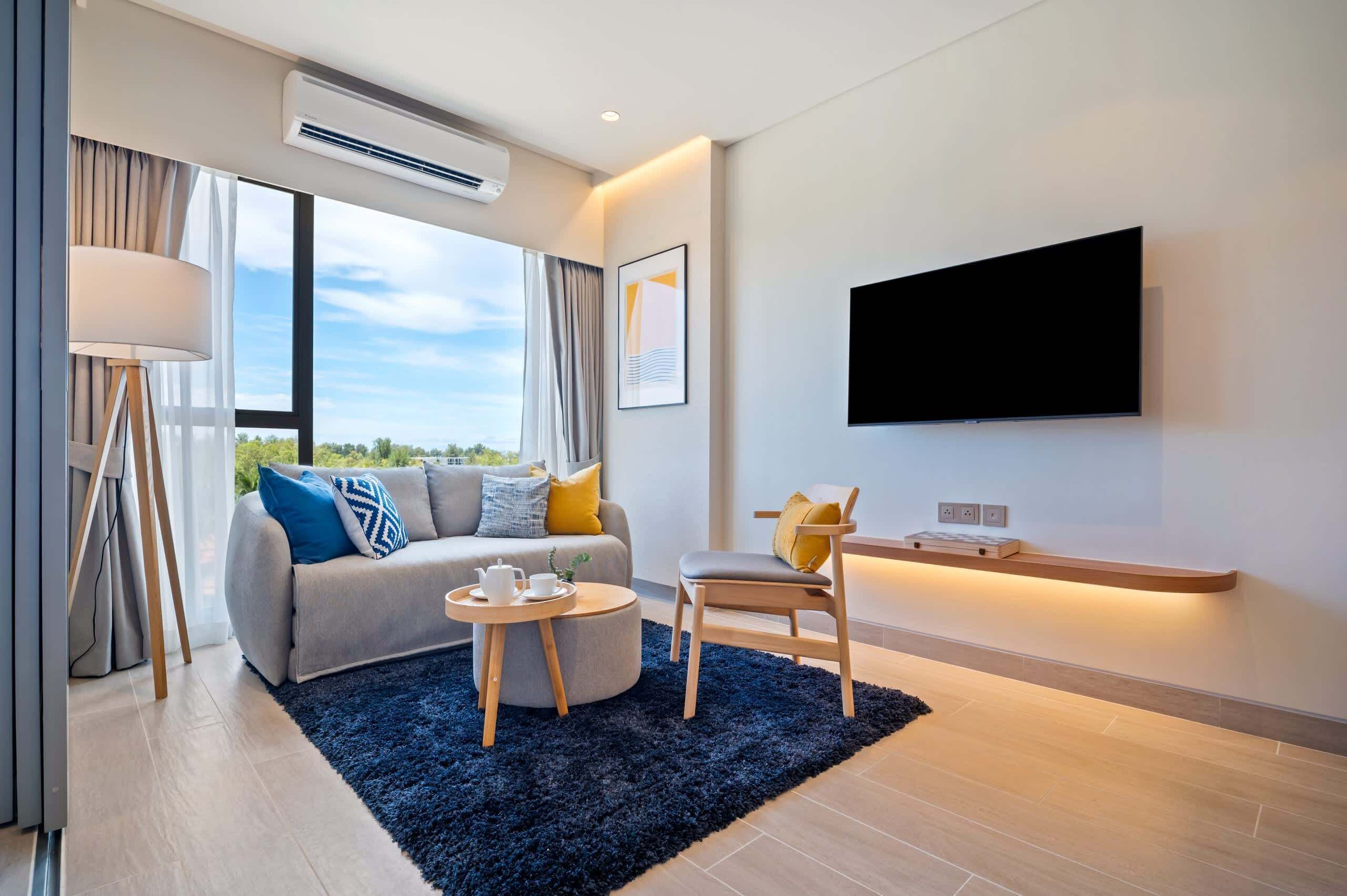 A modern living room with a sofa, a chair, a small round table with a teapot, a large window with curtains, and a flat-screen TV mounted on the wall.