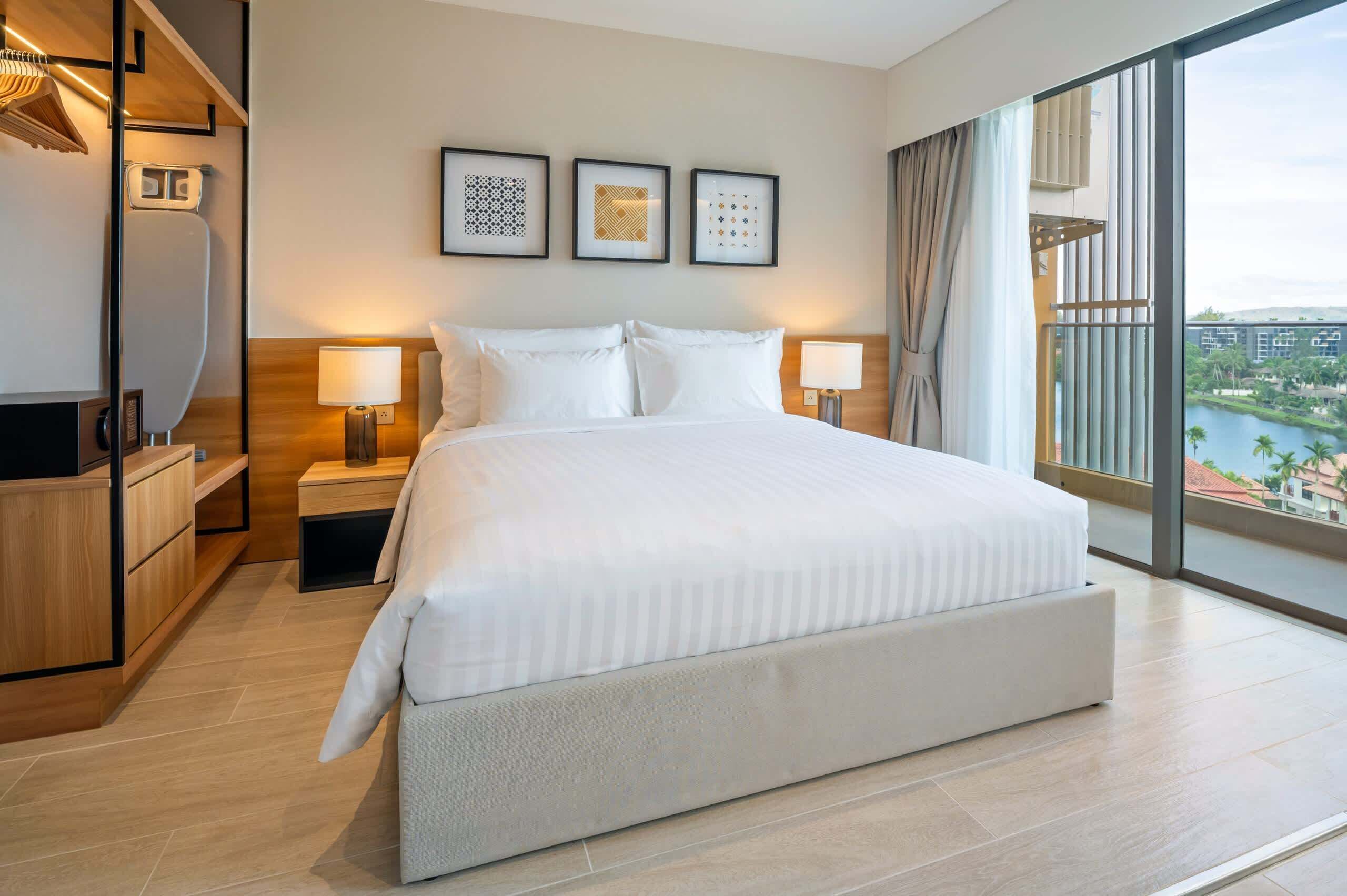 Une chambre d’hôtel moderne avec un grand lit, des lampes de chevet, une armoire et une vue sur le paysage urbain grâce à de grandes fenêtres.
