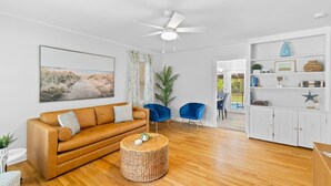 A modern living room with a brown leather sofa, a round wicker ottoman, a ceiling fan, and a view into a dining area with a pool. - 3BR Pool Home | Free Parking | Near Airport + DT (North Charleston)