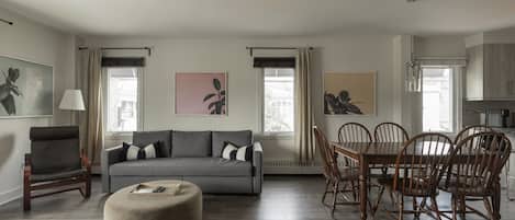 A modern living room with a grey sofa, wooden chairs, a round ottoman, and artwork on the walls.