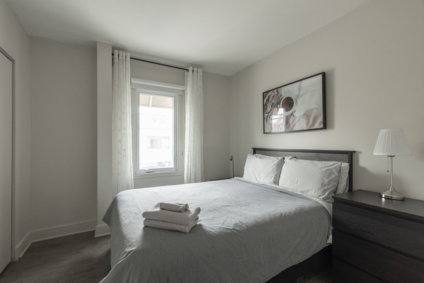 A minimalist bedroom with a bed, a bedside table with a lamp, a window with curtains, and a framed artwork on the wall.