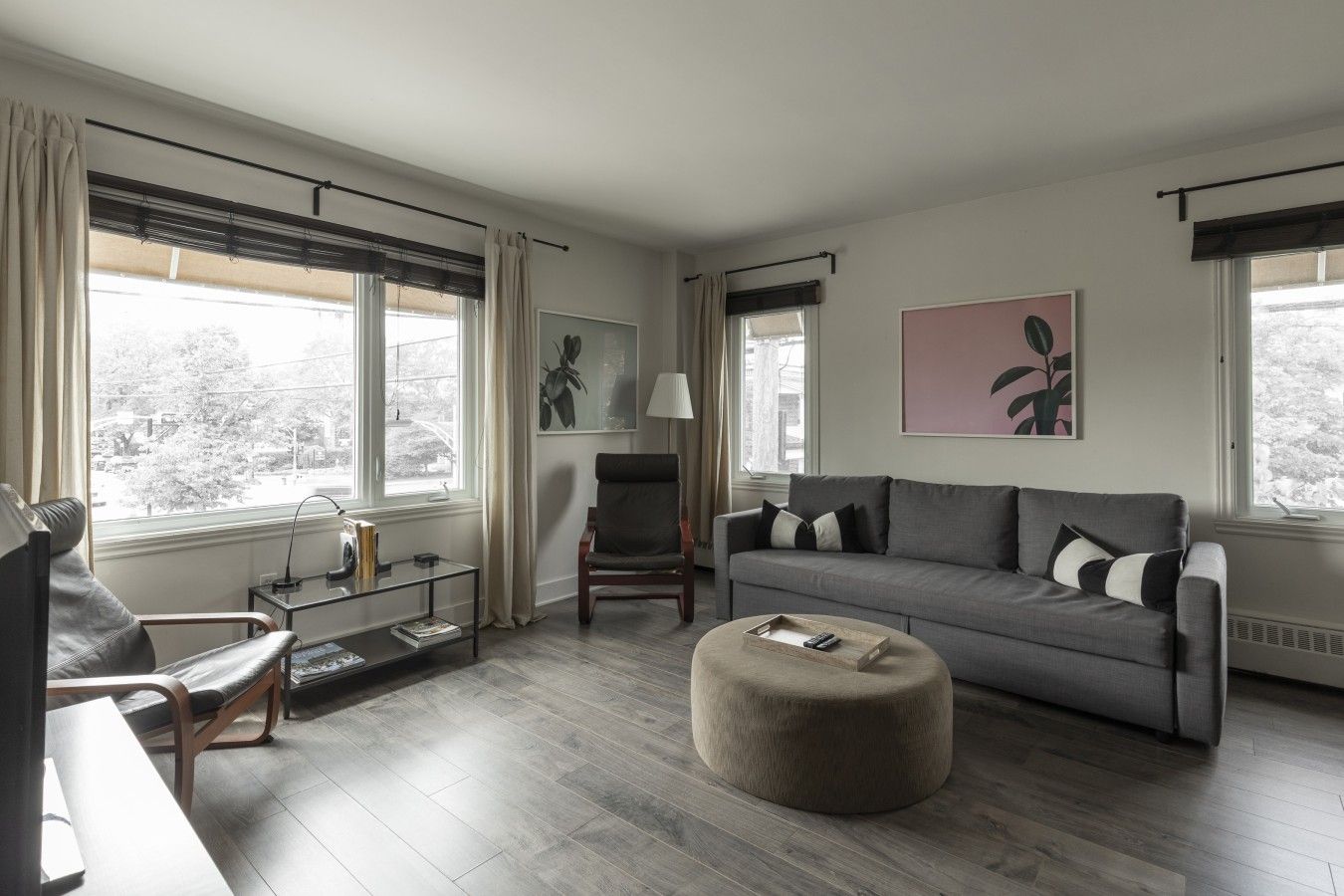 Un salon moderne avec un canapé gris, un pouf rond, une table basse en verre et un fauteuil. Il y a deux fenêtres avec des rideaux, une lampe et des œuvres d’art accrochées aux murs.