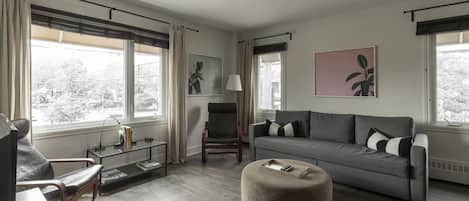 A modern living room with a grey sofa, a round ottoman, a glass coffee table, and a chair. There are two windows with curtains, a lamp, and artwork on the walls.