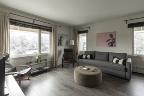 Moderni olohuone, jossa on harmaa sohva, pyöreä rahi, lasinen sohvapöytä ja tuoli. Huoneessa on kaksi ikkunaa, joissa on verhot, sekä lamppu ja taideteoksia seinillä.